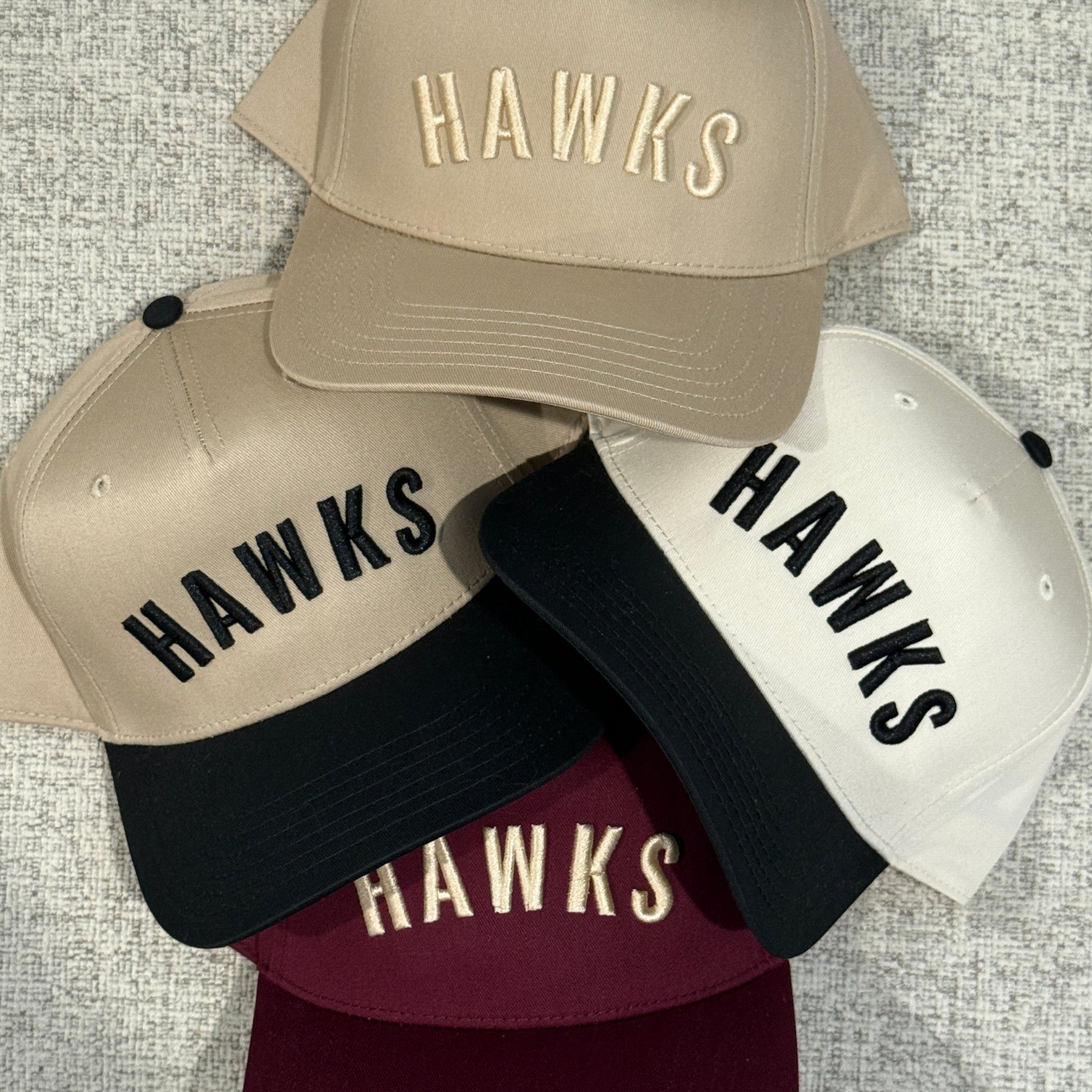 Four caps with 'HAWKS' branding in different colors on a textured surface.