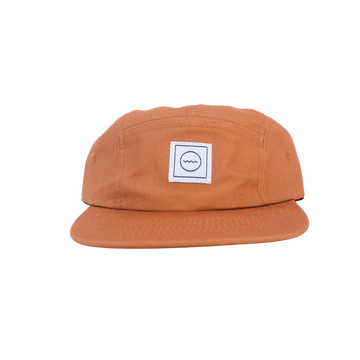 A brown cotton five-panel hat with an adjustable velcro back and fabric eyelets on the side panels, featuring a white square logo with a symbol in the center.