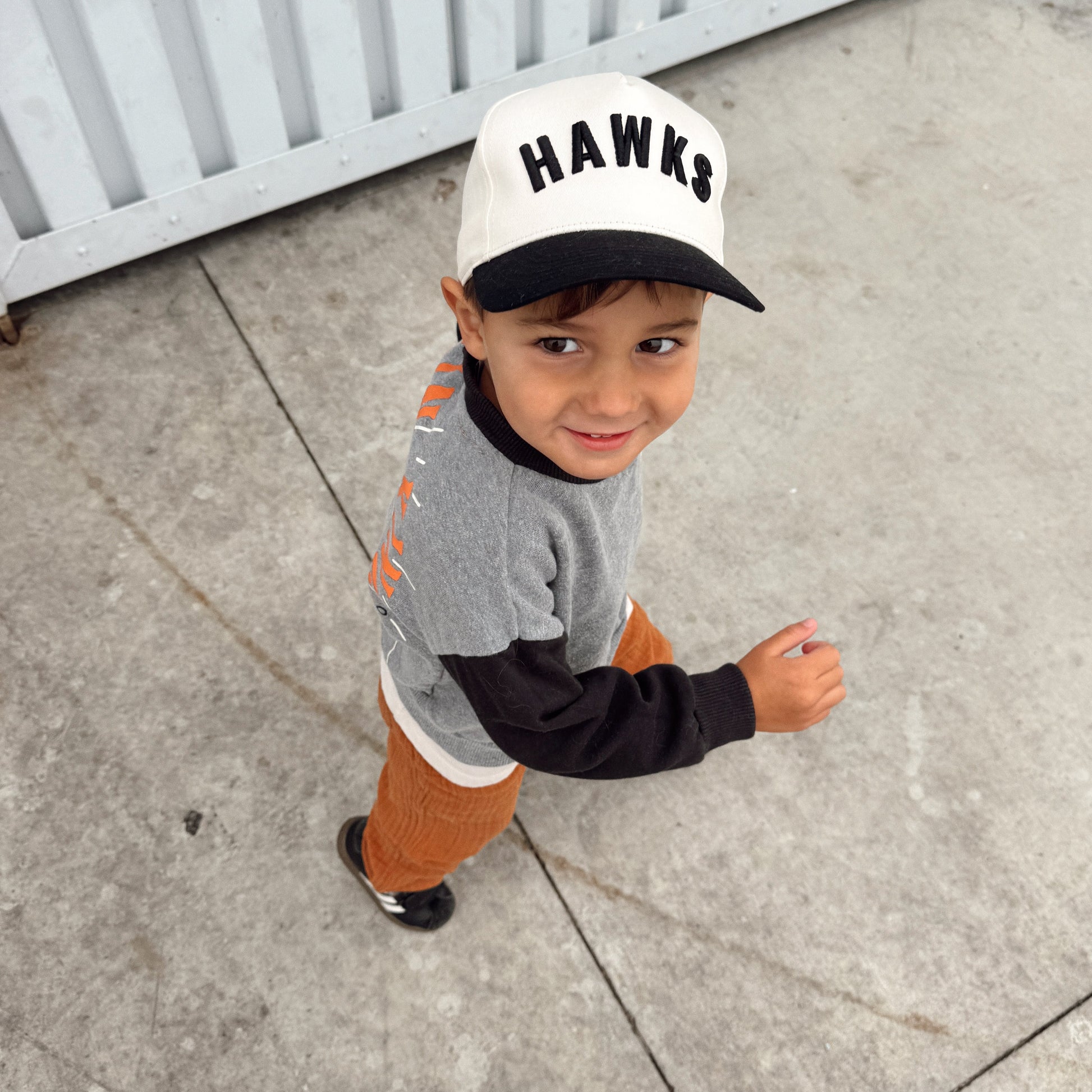A young boy wearing a white and black baseball cap with 'HAWKS' printed on the front, a gray and black long-sleeve shirt, and orange pants, standing on a concrete surface in front of a white corrugated metal wall.