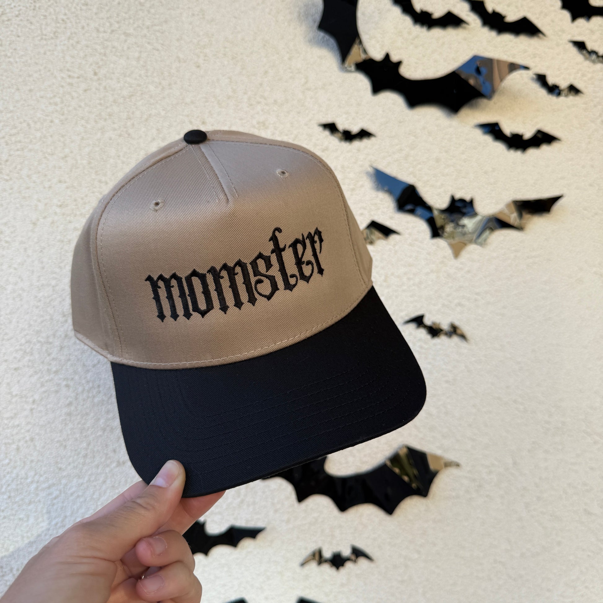Two-tone cap with 'momster' text held by a hand against a textured white background with black bat shapes.
