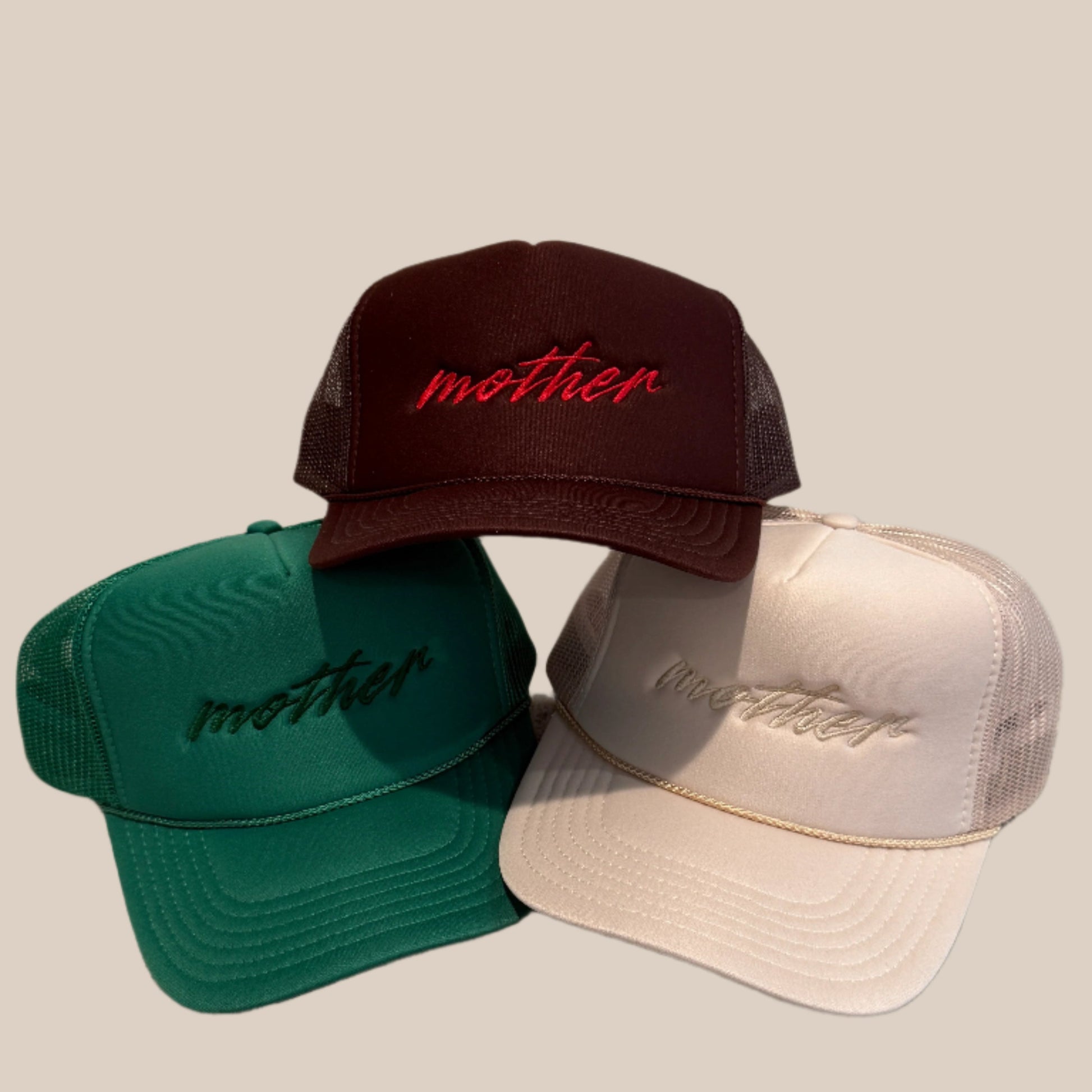 Three baseball caps in green, brown, and beige with 'mother' embroidered on them, placed on a textured surface.