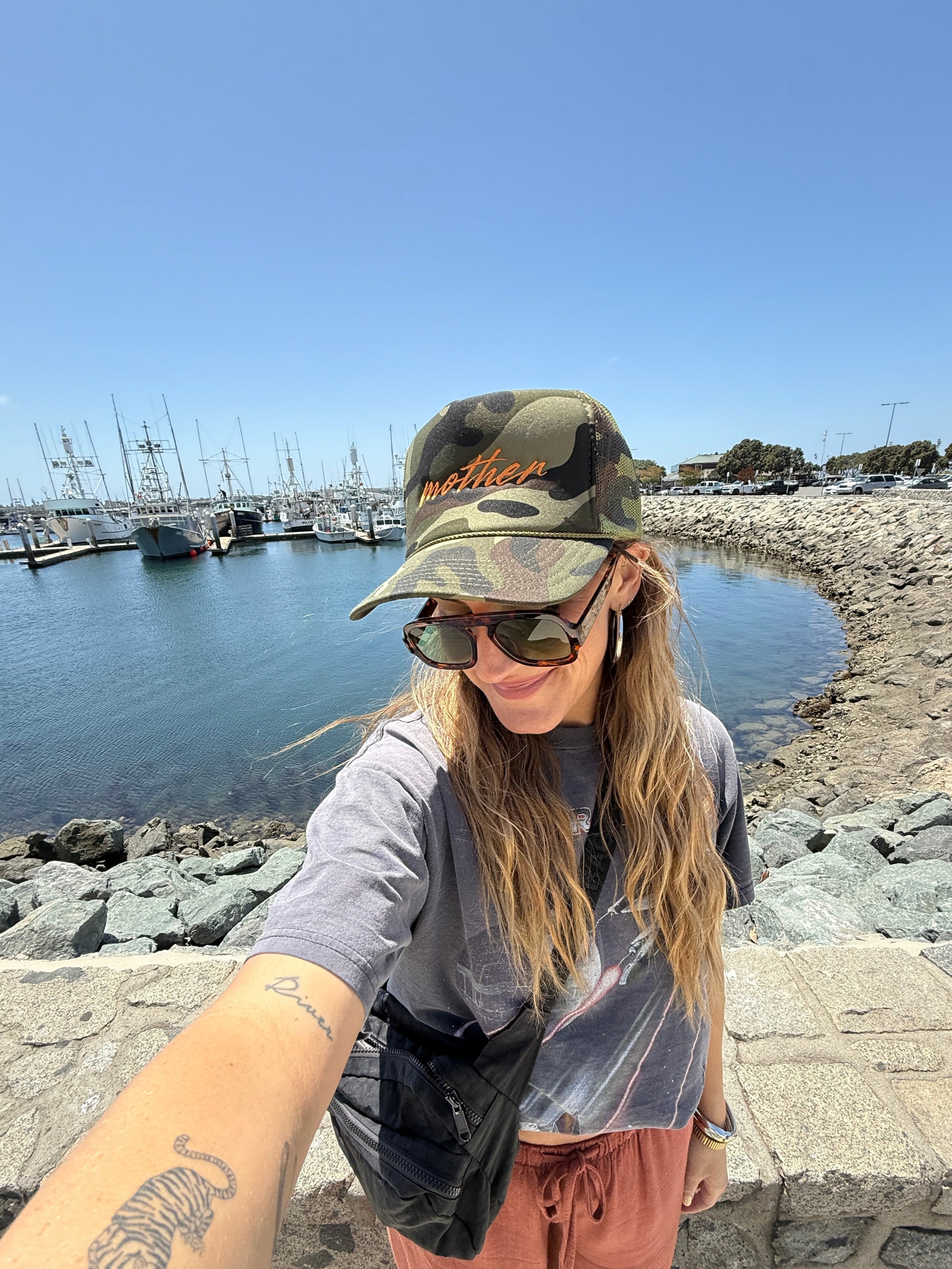 Person taking a selfie in a camo trucker hat with embroidered text by a waterfront with a clear blue sky